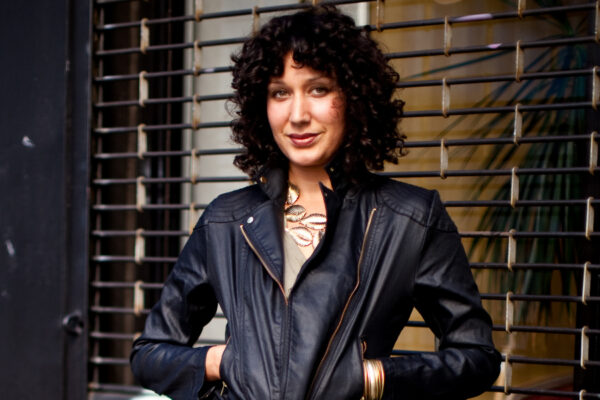 woman with dark curly hair wearing a black leather jacket standing in front of a brown brick wall